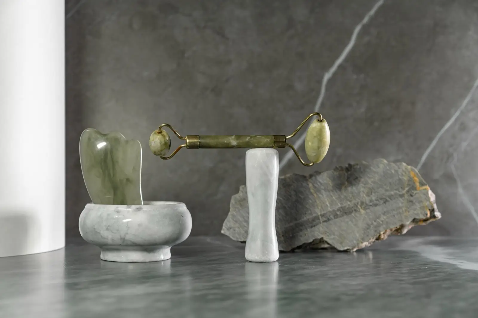 Jade-Roller, Gua-Sha-Stein und Marmorschale auf einer grauen Steinplatte