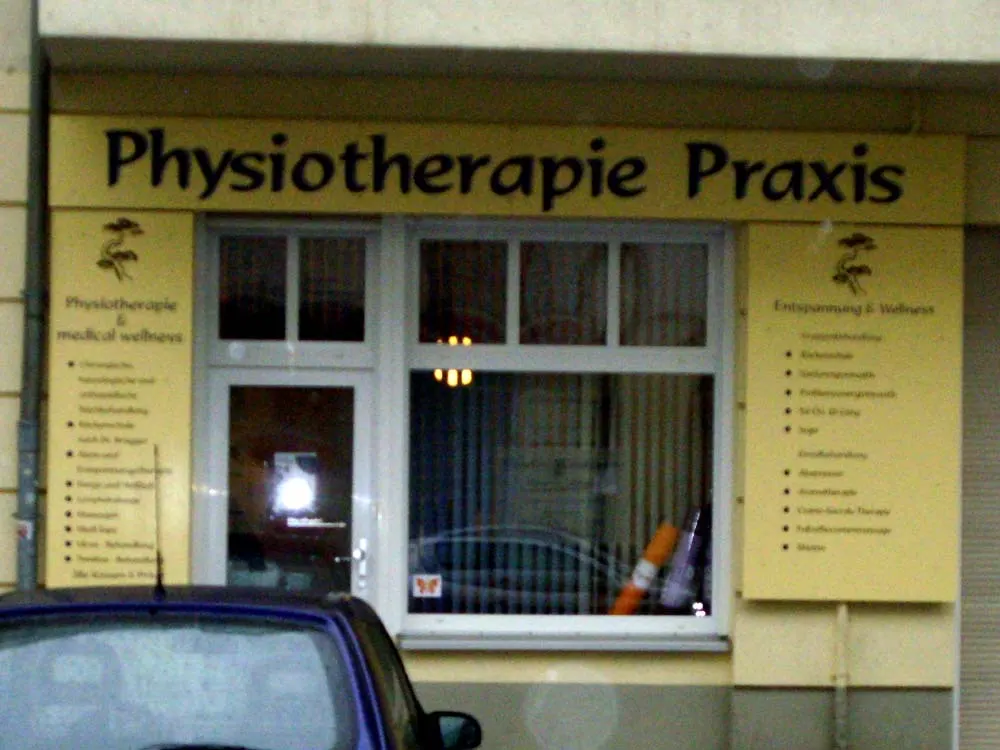 Gelbes Praxisschild mit der Aufschrift Physiotherapie Praxis über einem Schaufenster mit Leistungsübersicht