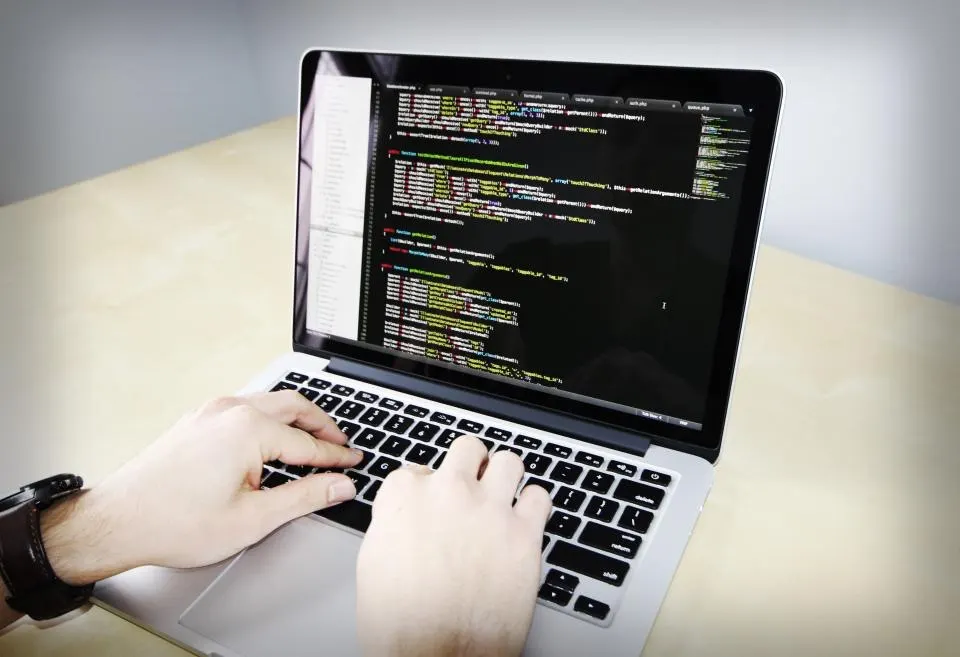 Hände tippen auf einem Laptop, auf dem Bildschirm ist Programmcode in einem Editor zu sehen
